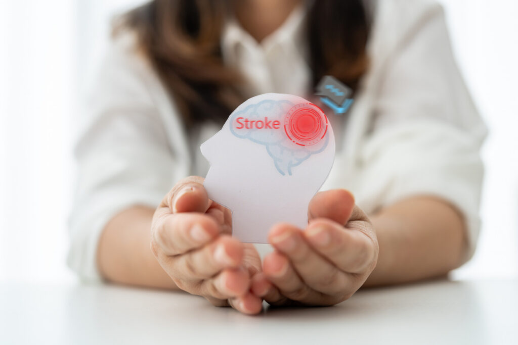 Woman hands holding paper head profile with red brain stroke text and warning icon, neurological disease awareness, brain attack symptom, medical healthcare and critical illness insurance concept